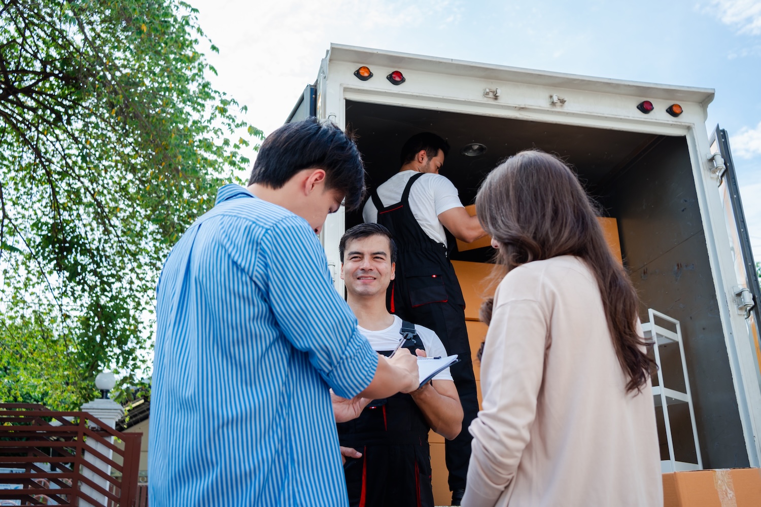 Asian Couple check while unloading boxes and furniture from a pickup truck to a new house with service cargo two men movers worker in uniform lifting boxes. concept of Home moving and delivery. Home Maintenance and Moving Services in Westminster
